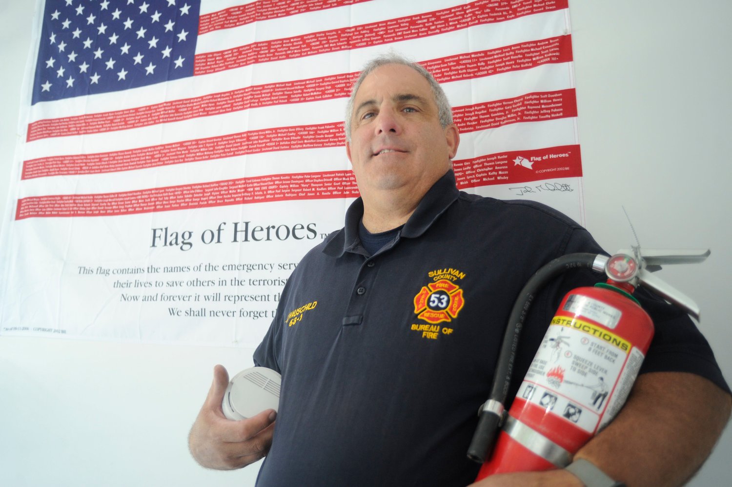 John Hauschild, Sullivan County fire coordinator and Emergency Services Training Center coordinator, proudly stands in front of the center’s “Flag of Heroes,” which lists the names of the emergency services personnel who gave their lives to save others on September 11, 2001...He is pictured holding a smoke detector and a fire extinguisher, two key elements of fire prevention...“We will always remember and never forget them... the sacrifices that were made by all,” said Hauschild of the roster of heroes.